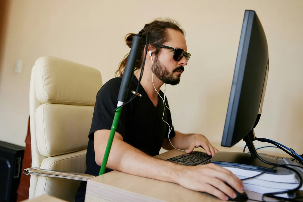 Junger Mann mit Sonnenbrille, Kopfhörern und Blindenstock am Arbeitsplatz