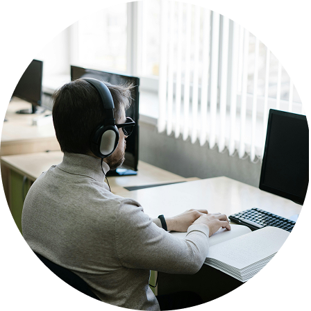 Person mit Kopfhörern sitzt an einem Schreibtisch vor zwei Monitoren und einem aufgeschlagenen Buch.